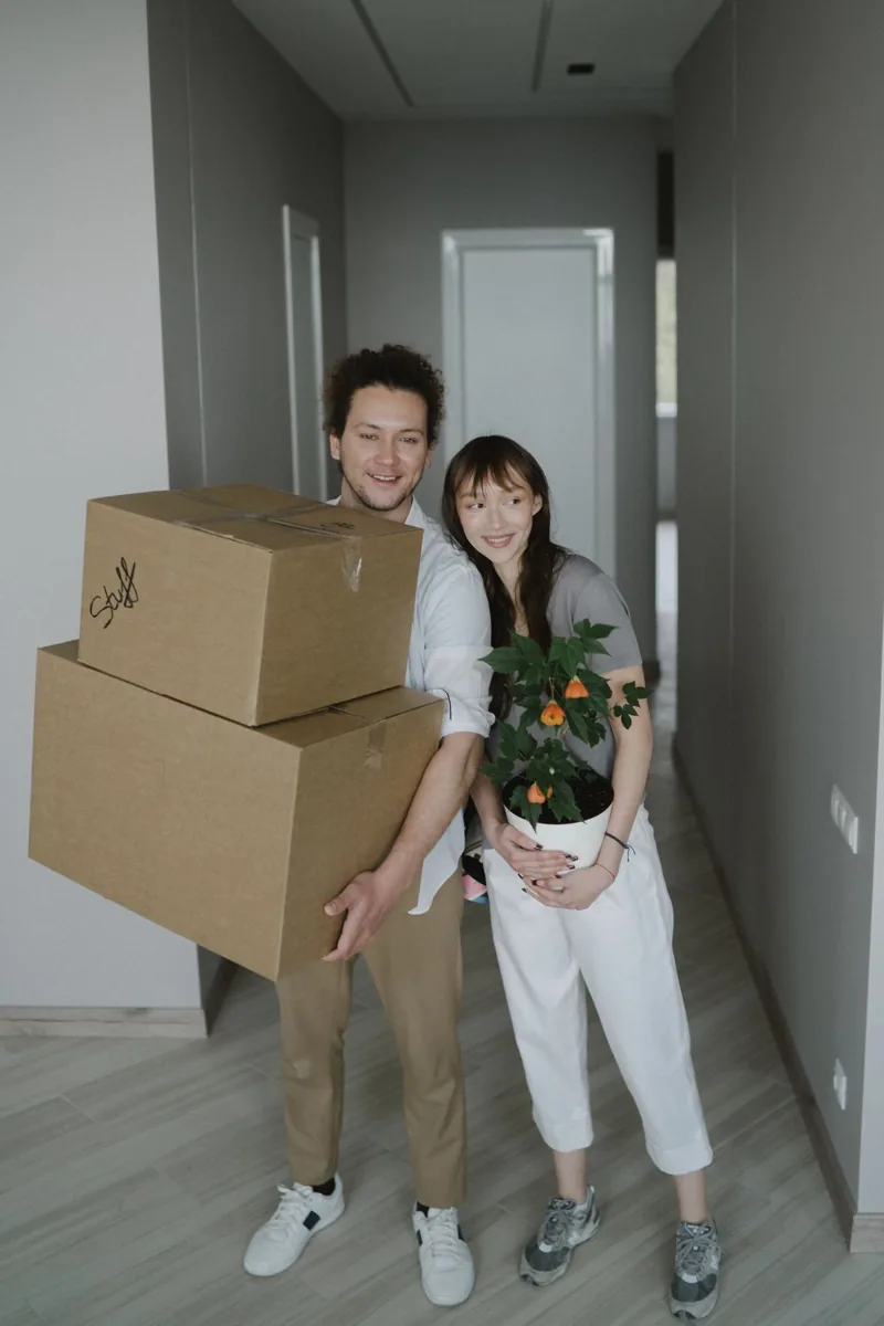 A couple carrying moving boxes and a plant into a home